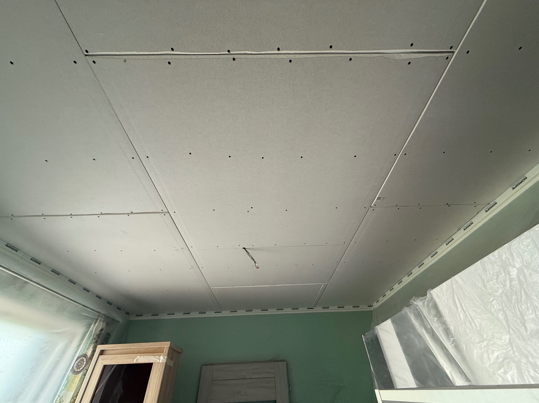 Montaż płyt karton-gips na suficie z widocznymi łączeniami i punktami mocowania. Częściowo zabezpieczone ściany folią malarską. Widoczne przewody elektryczne.