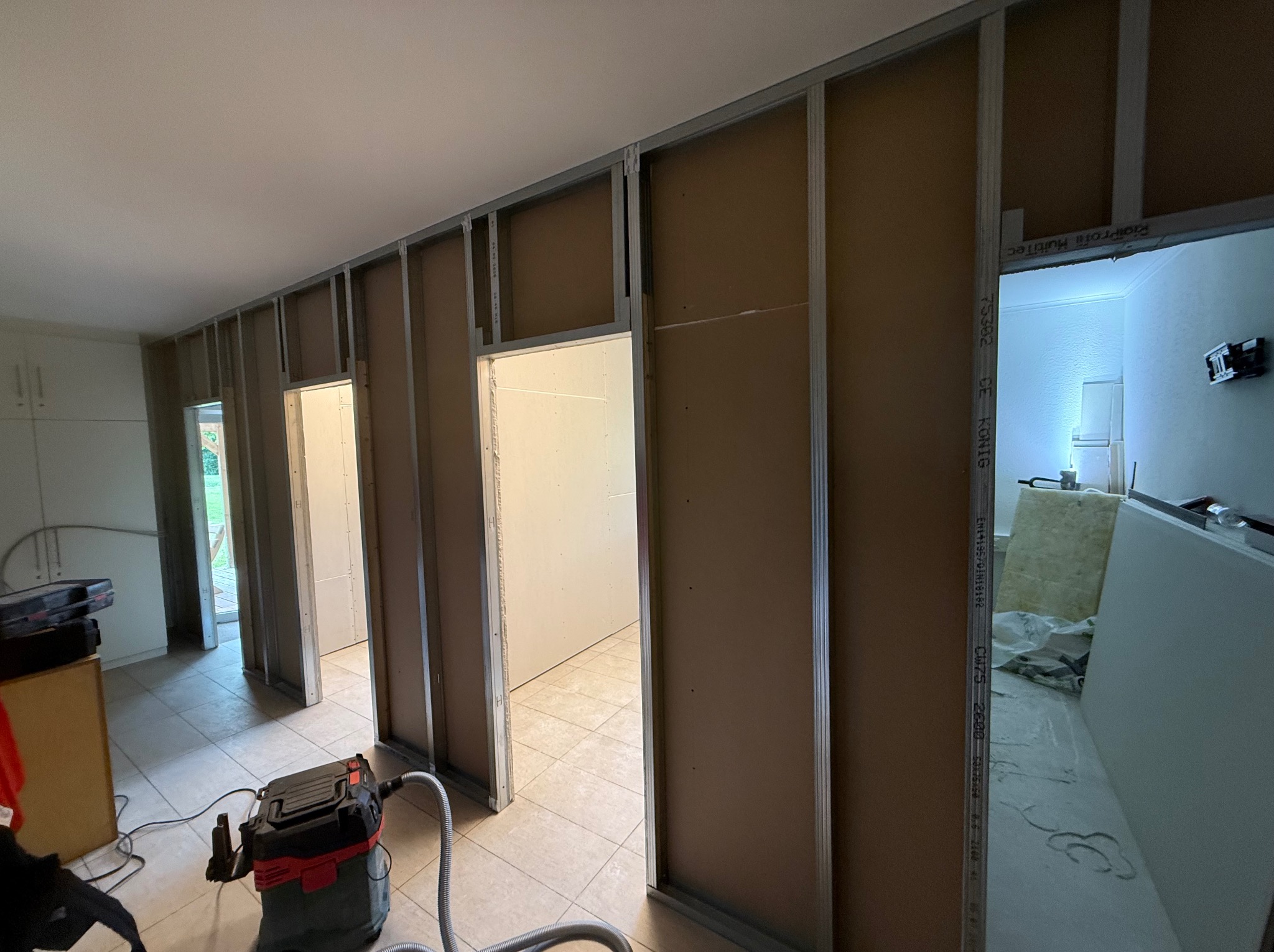 Montaż ścian działowych z płyty karton-gips na metalowym stelażu w trakcie prac wykończeniowych. Widoczny odkurzacz przemysłowy i fragment podłogi z płytek.