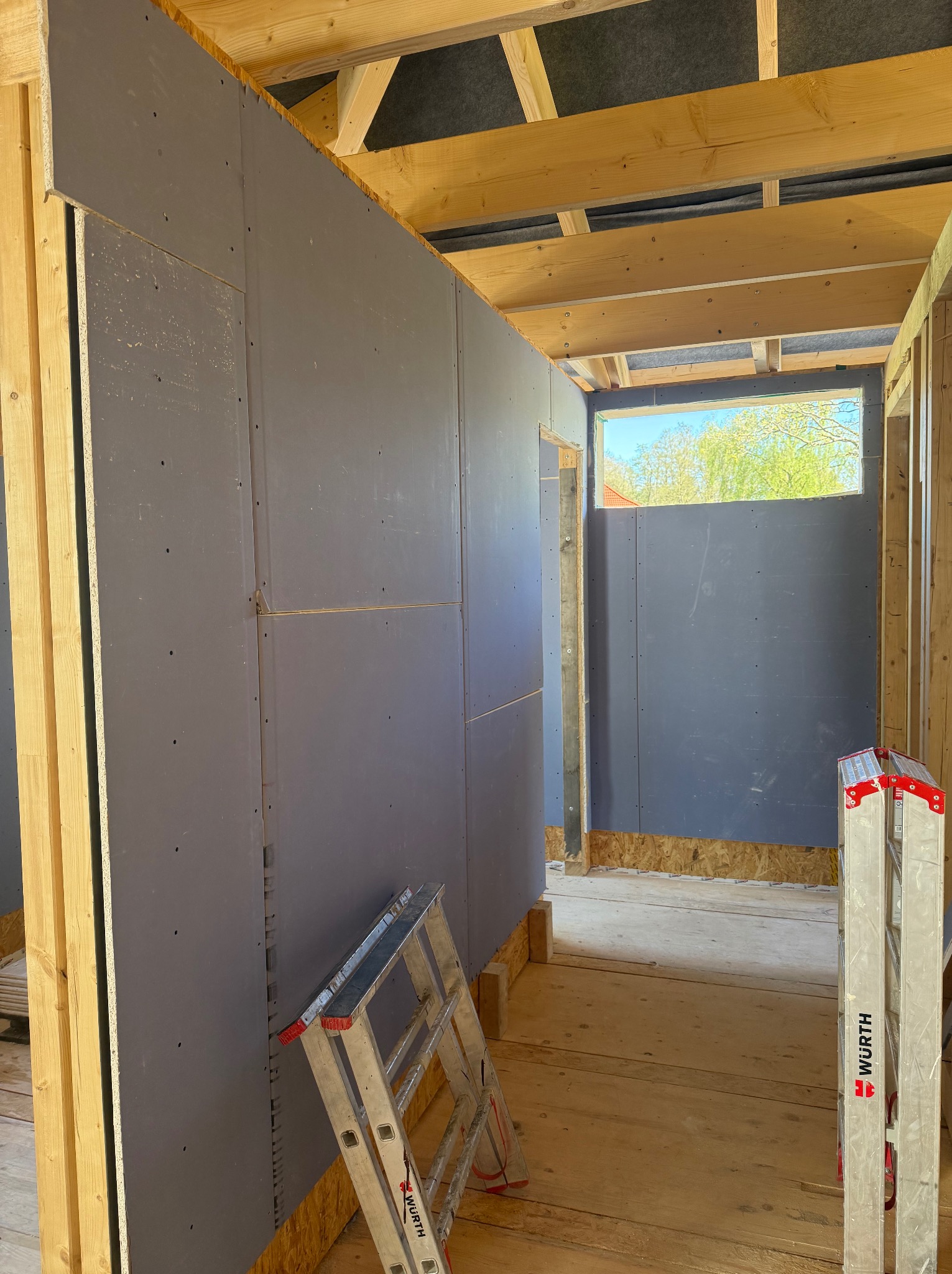 Wnętrze w trakcie prac: szare płyty karton-gips na drewnianej konstrukcji, widoczne fragmenty sufitu i podłogi. Dwie drabiny opierają się o ścianę. Otwór okienny z widokiem na zieleń.