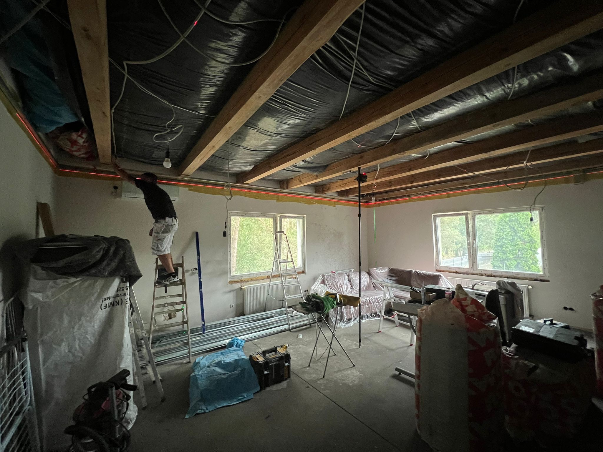 Montaż sufitu podwieszanego: mężczyzna na drabinie, laser wyznacza poziom, widoczne profile i folia. Remont w trakcie, okna z widokiem na zewnątrz.