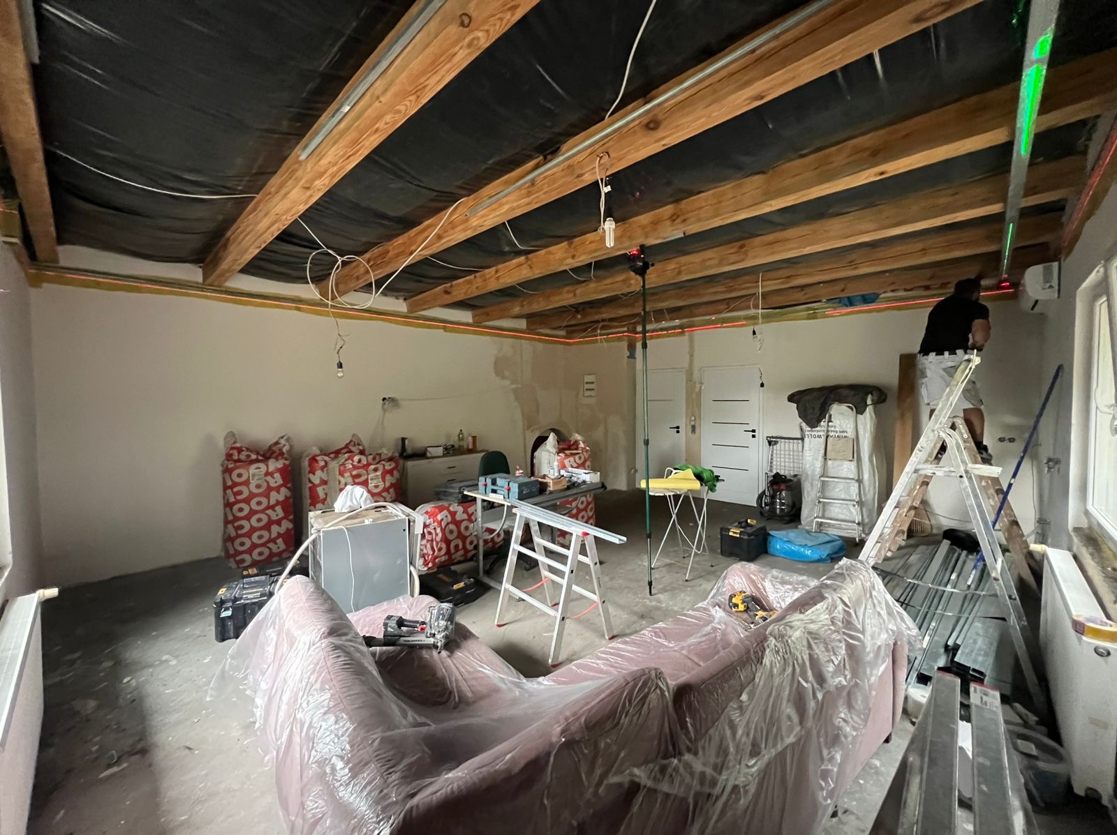 Remont wnętrza: widoczne belki stropowe z czarną folią, laserowy poziom, worki z wełną mineralną i narzędzia. Kanapa zabezpieczona folią malarską.