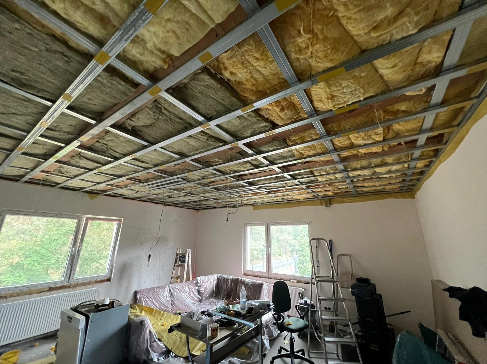 Sufit podwieszany z izolacją z wełny mineralnej, widoczny stelaż metalowy. Pokój w trakcie prac wykończeniowych, zabezpieczone meble folią malarską.