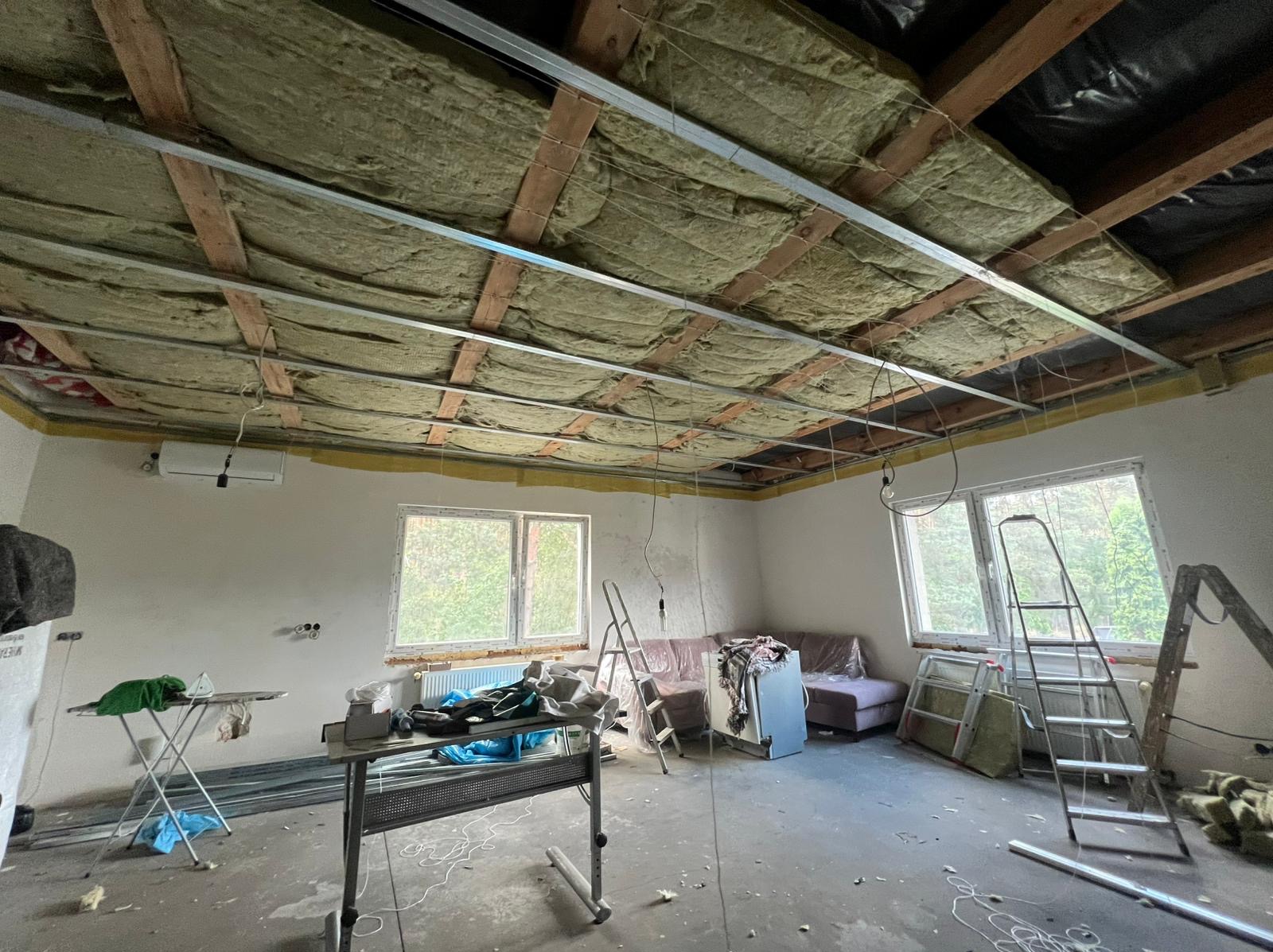 Montaż izolacji stropu wełną mineralną i metalowego stelaża w trakcie remontu. Widoczne okna, drabiny i narzędzia w pomieszczeniu.