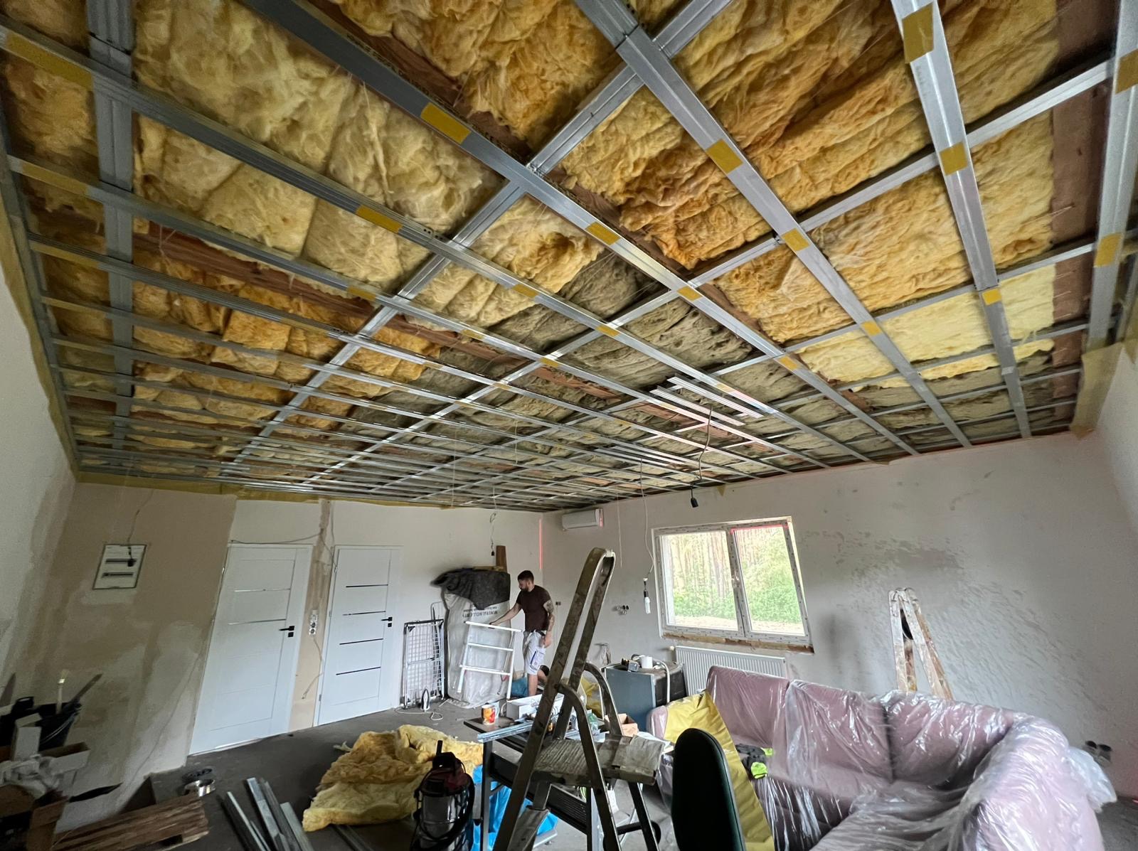 Montaż sufitu podwieszanego z izolacją z wełny mineralnej. Widoczna metalowa konstrukcja i fragment pomieszczenia w trakcie prac wykończeniowych.