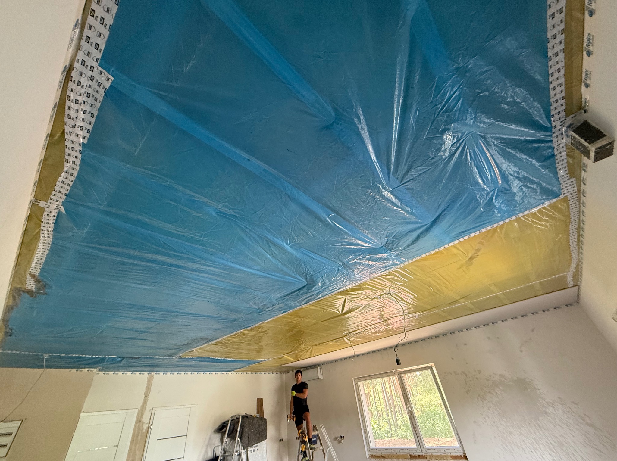 Montaż izolacji na suficie z folią w kolorach niebieskim i żółtym, widok z dołu na pracownika na drabinie w trakcie prac wykończeniowych w pomieszczeniu z oknem.