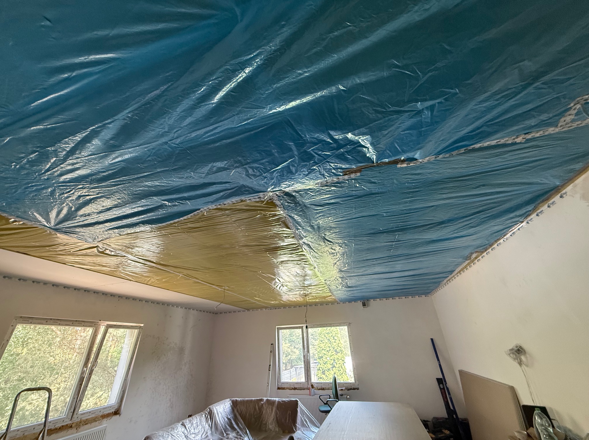 Pokój w trakcie zabezpieczania folią malarską sufitu (częściowo żółtą, częściowo niebieską) przed pracami remontowymi. Widoczne okna i meble przykryte folią.