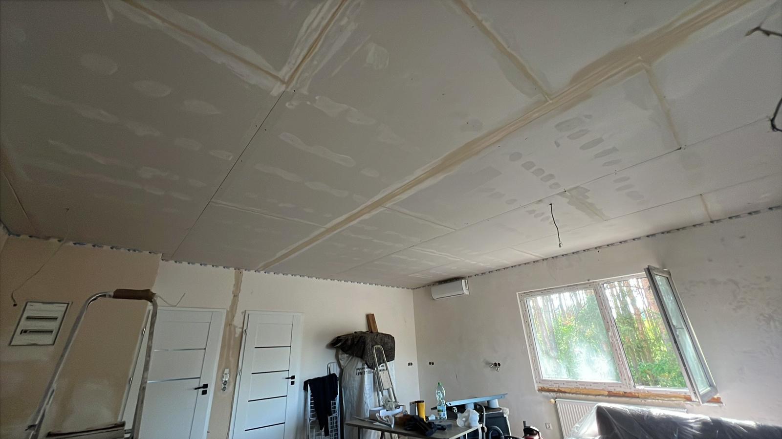 Sufit z płyt gipsowo-kartonowych w trakcie montażu, widoczne łączenia płyt, drabina i narzędzia w tle. Wnętrze w trakcie remontu, okno z widokiem na zieleń.