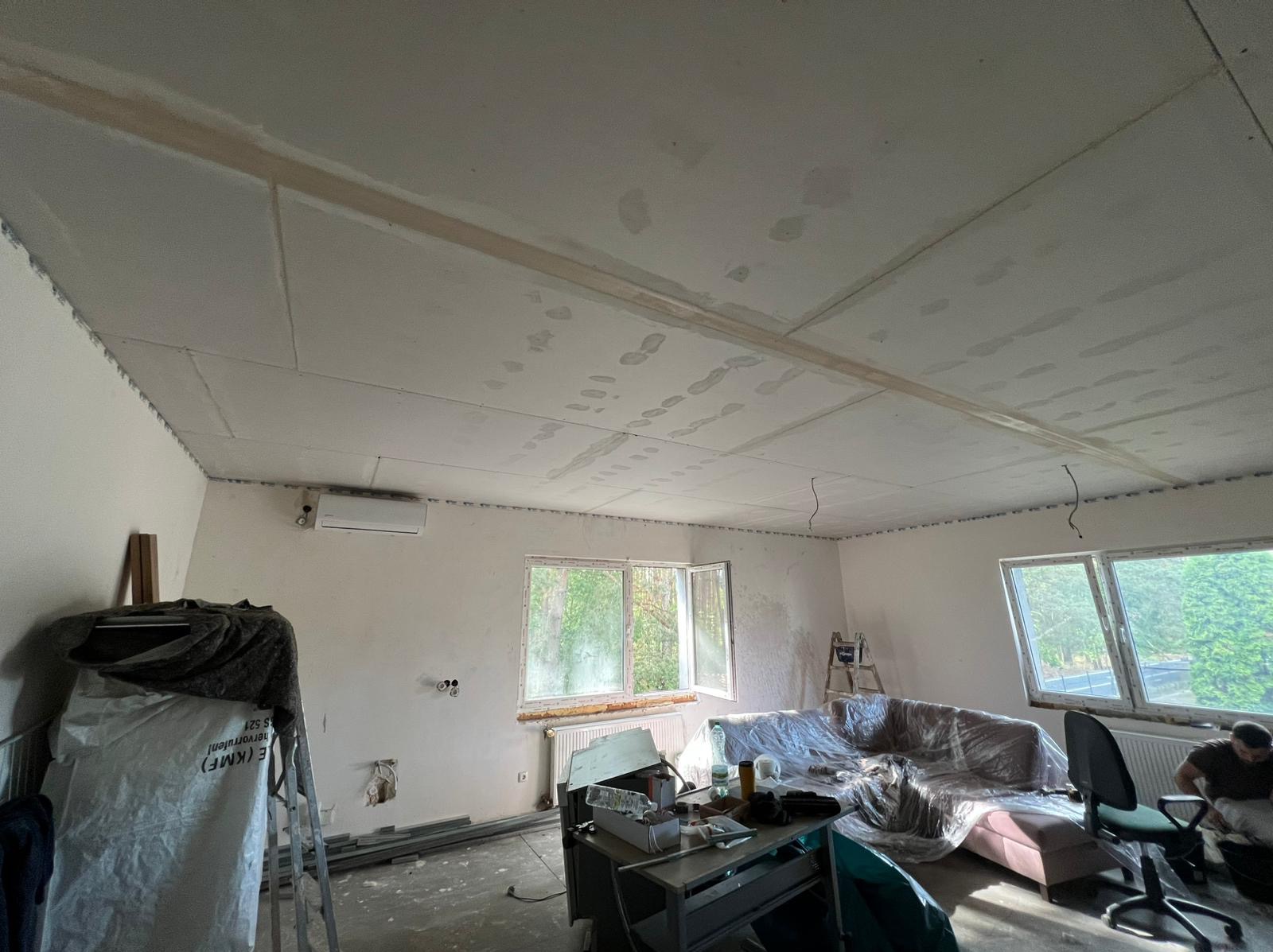 Pokój w trakcie remontu: zamontowane płyty gipsowe na suficie, widoczne łączenia i szpachlowane miejsca. Okna z widokiem na zieleń, meble przykryte folią.