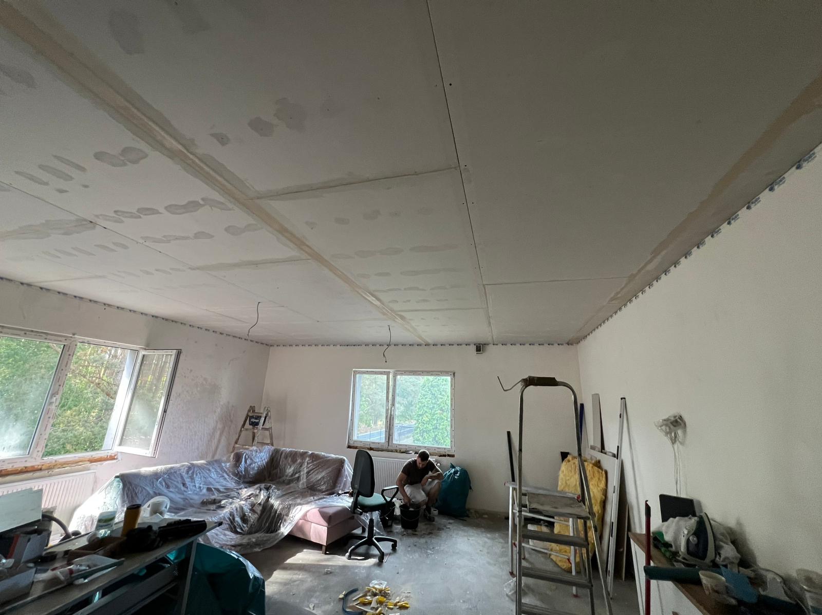 Montaż płyt gipsowo-kartonowych na suficie w trakcie remontu. Widoczne płyty, profile, drabina i osoba przygotowująca masę szpachlową. Meble zabezpieczone folią.