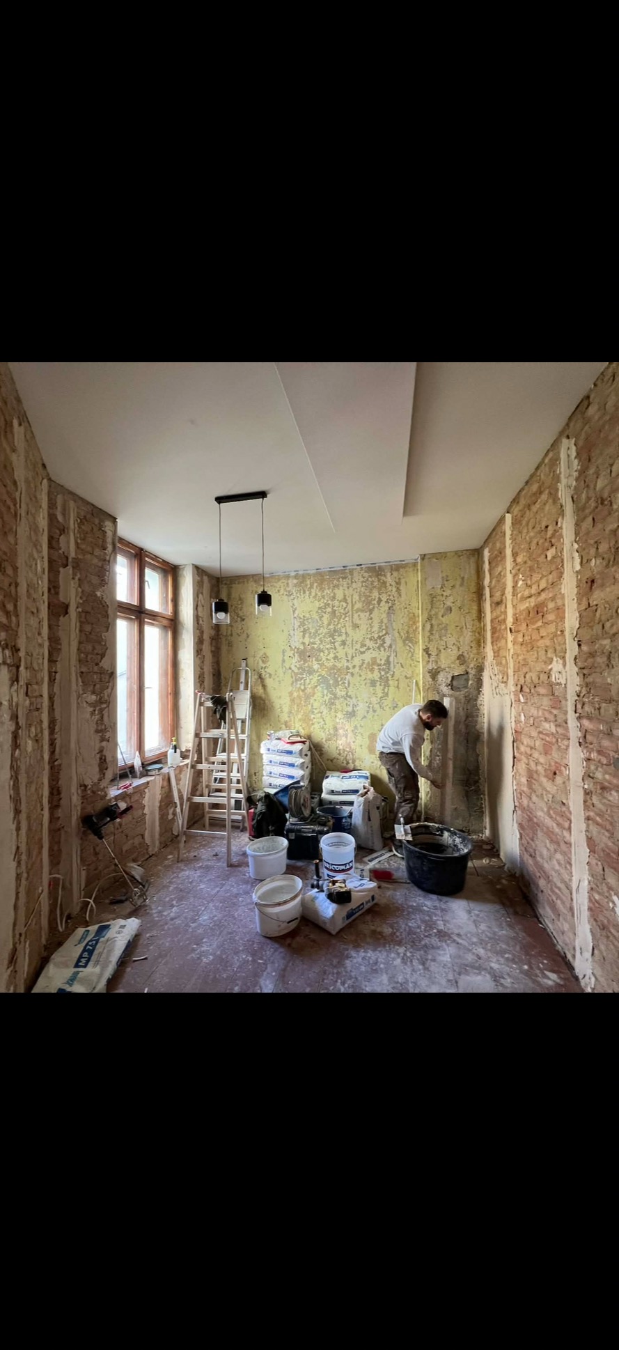 Remont pokoju: murarz nakłada gładź na ścianę z cegły, widoczne worki z zaprawą, drabina i lampa sufitowa. Częściowo otynkowany sufit.