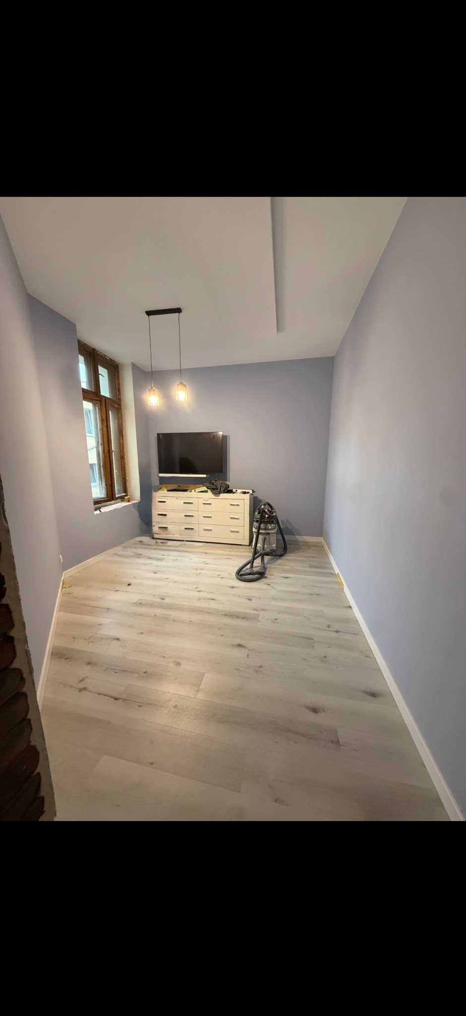 Pokój po remoncie: świeżo pomalowane ściany w odcieniach fioletu, jasne panele podłogowe, okno z drewnianą ramą i wiszące lampy. Widoczny odkurzacz przemysłowy.