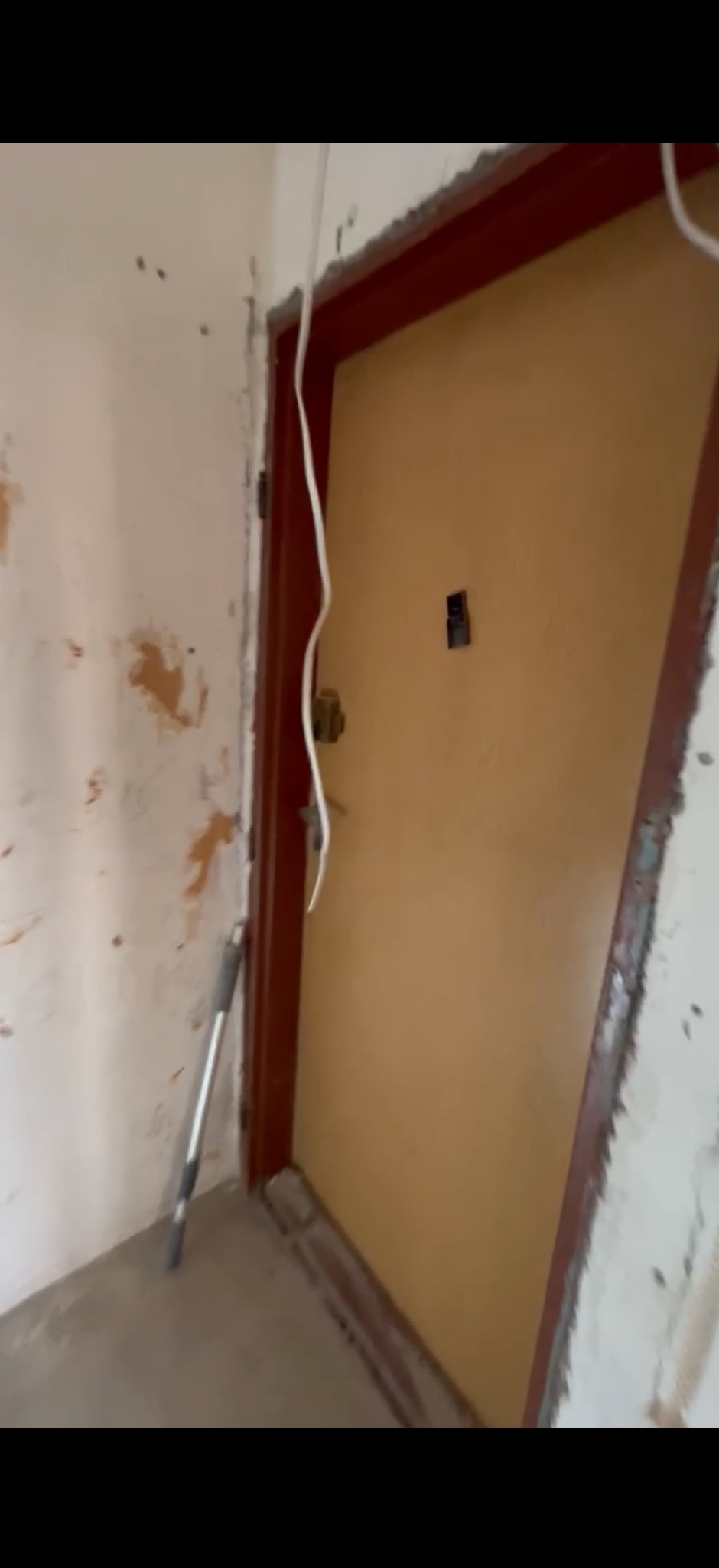 Ściana z uszkodzeniami i wystającymi kablami, obok ościeżnica drzwi z widocznymi śladami prac remontowych. W tle widoczne drzwi.