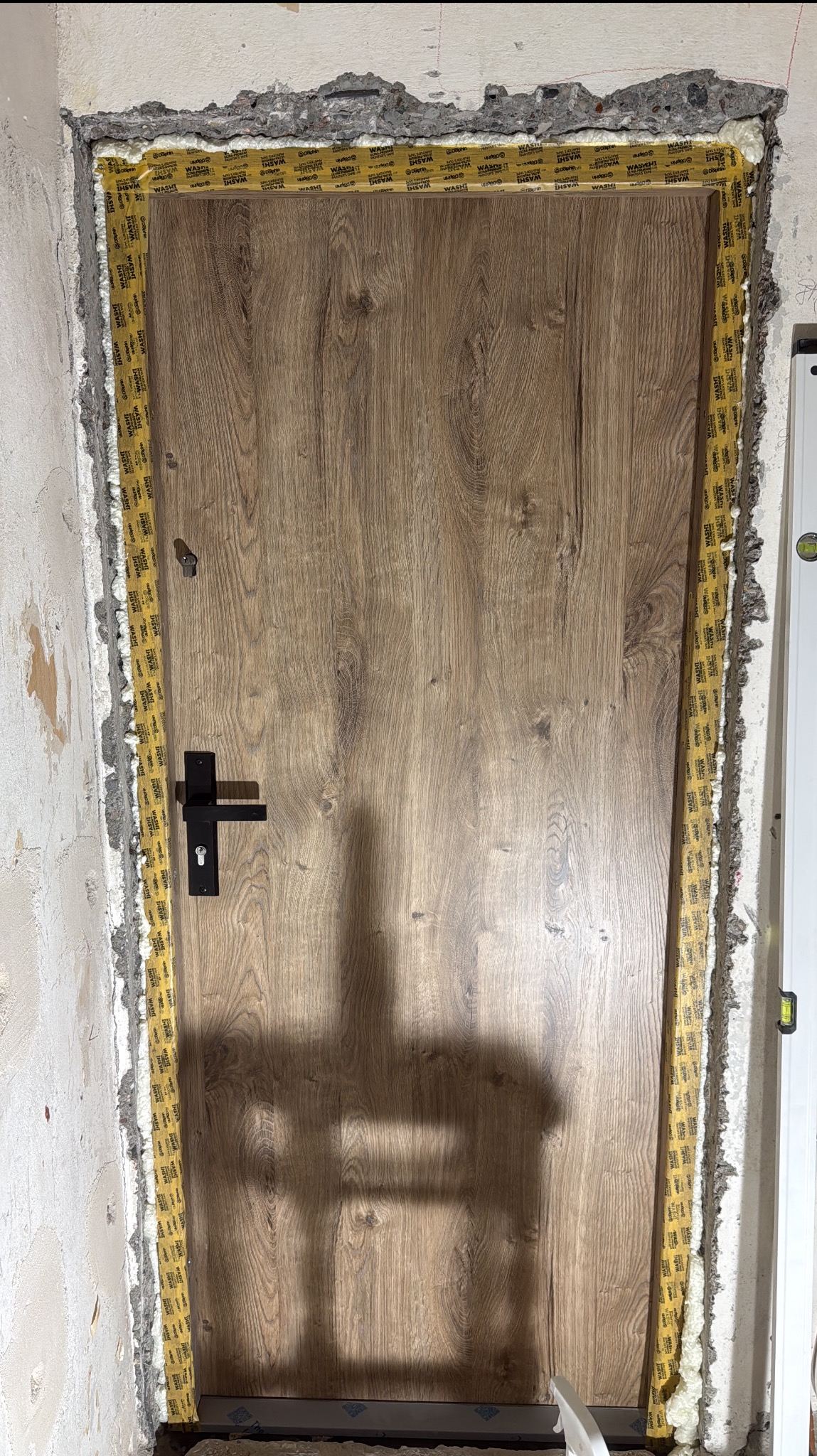 Montaż ościeżnicy drzwiowej z drewnopodobnym wykończeniem, zabezpieczonej taśmą malarską i pianką montażową, w surowym otworze ściany. Widoczny poziomica.