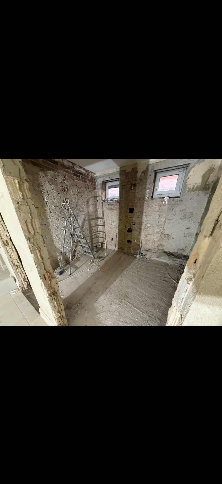 Wnętrze w trakcie remontu: odsłonięta cegła, częściowo otynkowane ściany, drabina, przygotowana podłoga. Widoczne okna i instalacje. Surowy stan przed wykończeniem.