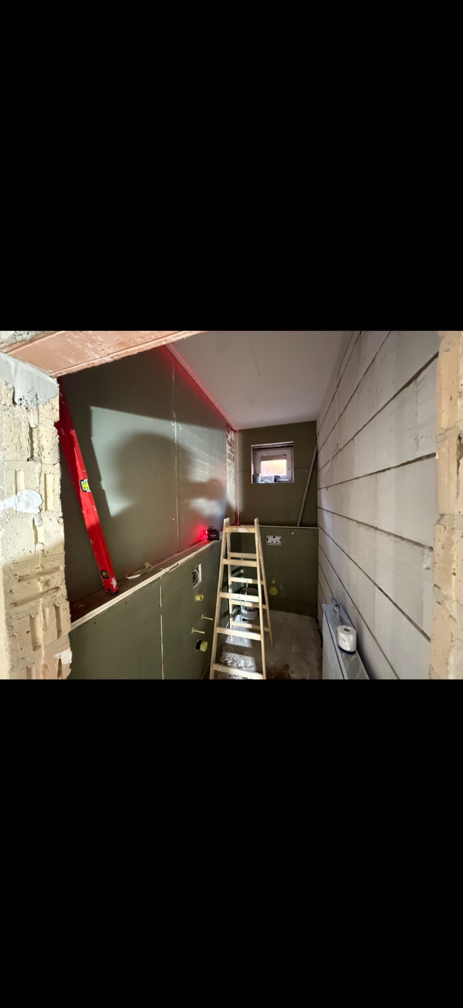 Wnętrze łazienki w trakcie remontu: ściany z płytami, poziomica laserowa, drabina. Widoczne instalacje i przygotowanie do dalszych prac wykończeniowych.