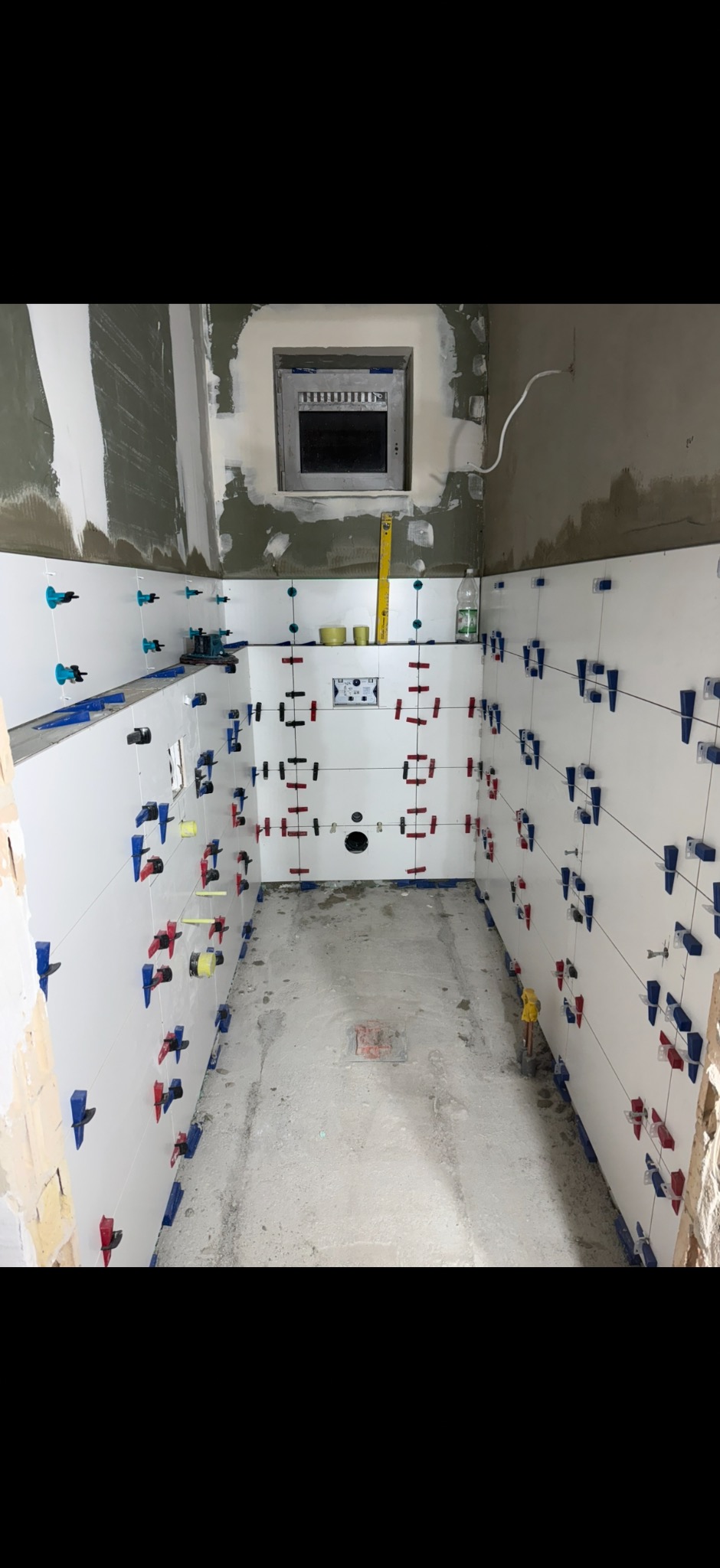 Białe płytki ścienne w trakcie montażu w łazience, z użyciem klipsów poziomujących w kolorach czerwonym, niebieskim i zielonym. Widoczna instalacja podtynkowa.