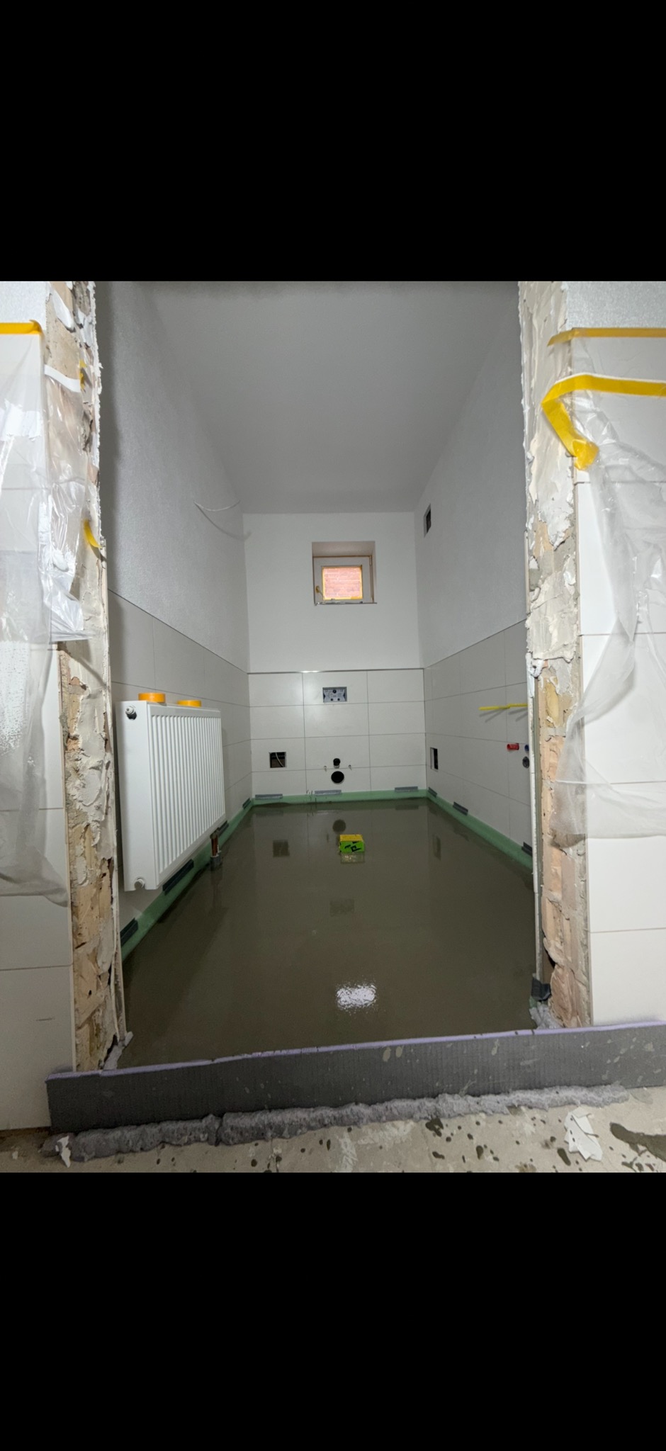 Wnętrze łazienki w trakcie wylewania posadzki samopoziomującej. Widoczne płytki na ścianach, grzejnik i przygotowane obrzeża. Prace wykończeniowe w toku.