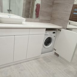 MobiliCraft KONRAD WÓJTOWICZ - Biała, nowoczesna szafka łazienkowa z umywalką nablatową i wbudowaną pralką, z otwartymi drzwiczkami po prawej stronie, na tle płytek imitujących kamień.