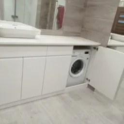 Biała, nowoczesna szafka łazienkowa z umywalką nablatową i wbudowaną pralką, z otwartymi drzwiczkami po prawej stronie, na tle płytek imitujących kamień.