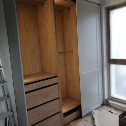 MobiliCraft KONRAD WÓJTOWICZ - Wnętrze szafy wnękowej z drewnianymi elementami i szufladami, widoczna drabina i kartony na podłodze, okno w tle.