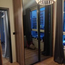 MobiliCraft KONRAD WÓJTOWICZ - Szafa przesuwna z lustrzanymi drzwiami w pokoju, widoczne odbicie okna z budynkiem i zasłony, drewniana podłoga i lampa sufitowa.
