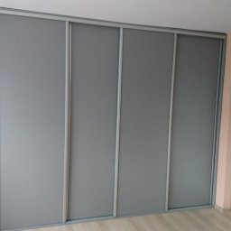 MobiliCraft KONRAD WÓJTOWICZ - Czterodrzwiowa szafa wnękowa z przesuwnymi, szarymi drzwiami w pokoju o ścianach w kolorze brzoskwiniowym, widoczna biała ościeżnica drzwiowa po prawej stronie.