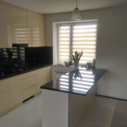 MobiliCraft KONRAD WÓJTOWICZ - Jasna kuchnia z beżowymi frontami, czarnym blatem i wyspą kuchenną, rolety w oknie, odbicia światła na blacie.