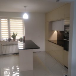 MobiliCraft KONRAD WÓJTOWICZ - Jasna kuchnia z czarnym blatem, oświetleniem punktowym i wyspą kuchenną, widok na okno z roletami i roślinami doniczkowymi.