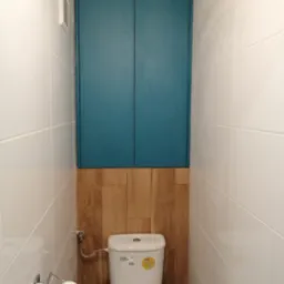 Wnętrze toalety z zabudowaną szafką w kolorze turkusowym nad deską sedesową, wykończoną drewnopodobnymi panelami, oraz białą glazurą na ścianach.