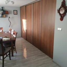 Długi rząd szaf wnękowych z przesuwnymi drzwiami o drewnianym wykończeniu w jadalni, widoczny stół z krzesłami i dekoracyjny zegar na ścianie.