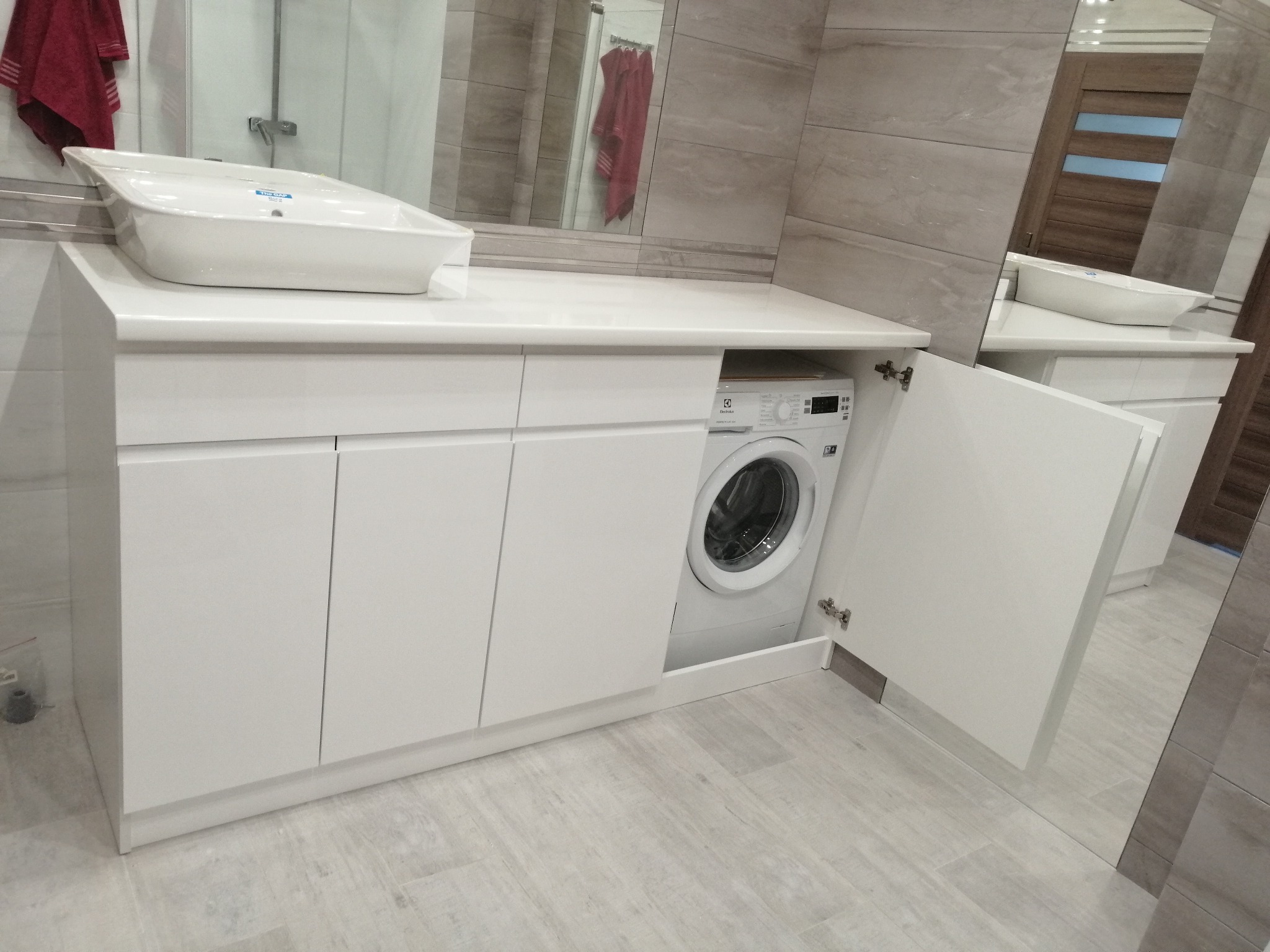 Biała, nowoczesna szafka łazienkowa z umywalką nablatową i wbudowaną pralką, z otwartymi drzwiczkami po prawej stronie, na tle płytek imitujących kamień.