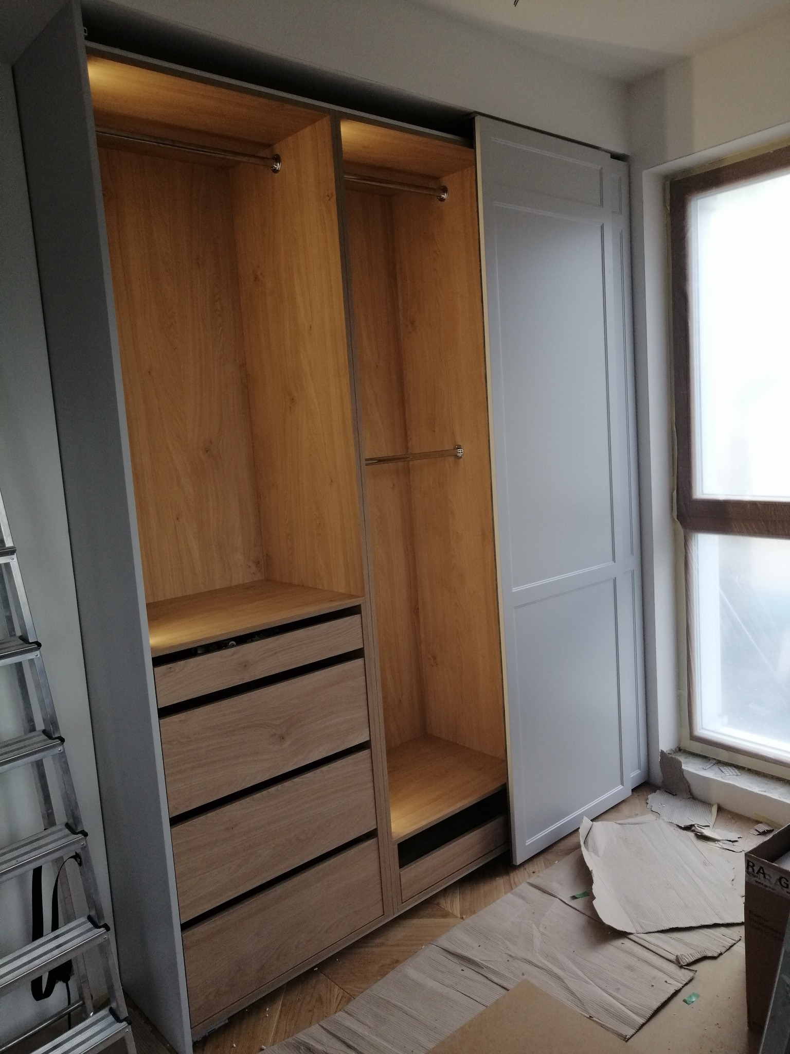 Wnętrze szafy wnękowej z drewnianymi elementami i szufladami, widoczna drabina i kartony na podłodze, okno w tle.