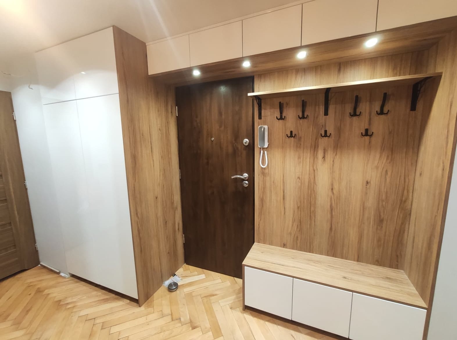 Przedpokój z zabudową: biała szafa przesuwna, panel ścienny z wieszakami i półką, oraz szafka na buty z siedziskiem w kolorze drewna, podłoga parkiet w jodełkę.