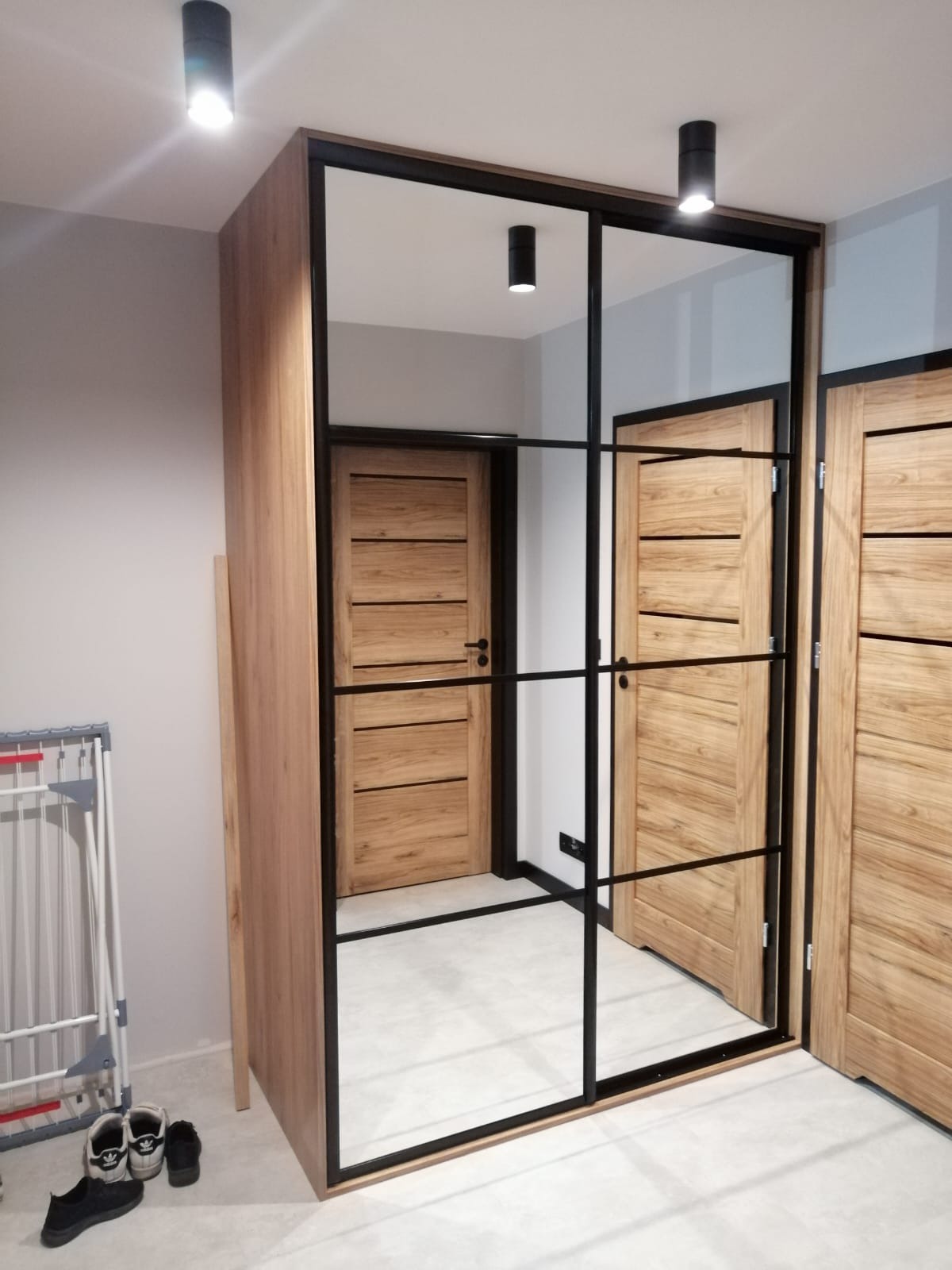 Szafa wnękowa z lustrem i czarnymi profilami w stylu loft, odbijająca drzwi z drewnianymi panelami, zrealizowana w Milejowie. Na podłodze widoczne buty i suszarka na pranie.