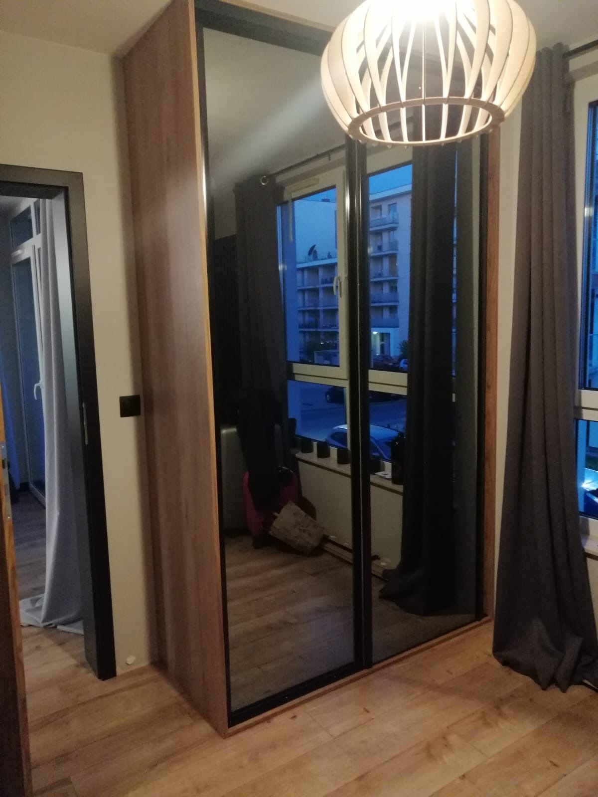 Szafa przesuwna z lustrzanymi drzwiami w pokoju, widoczne odbicie okna z budynkiem i zasłony, drewniana podłoga i lampa sufitowa.