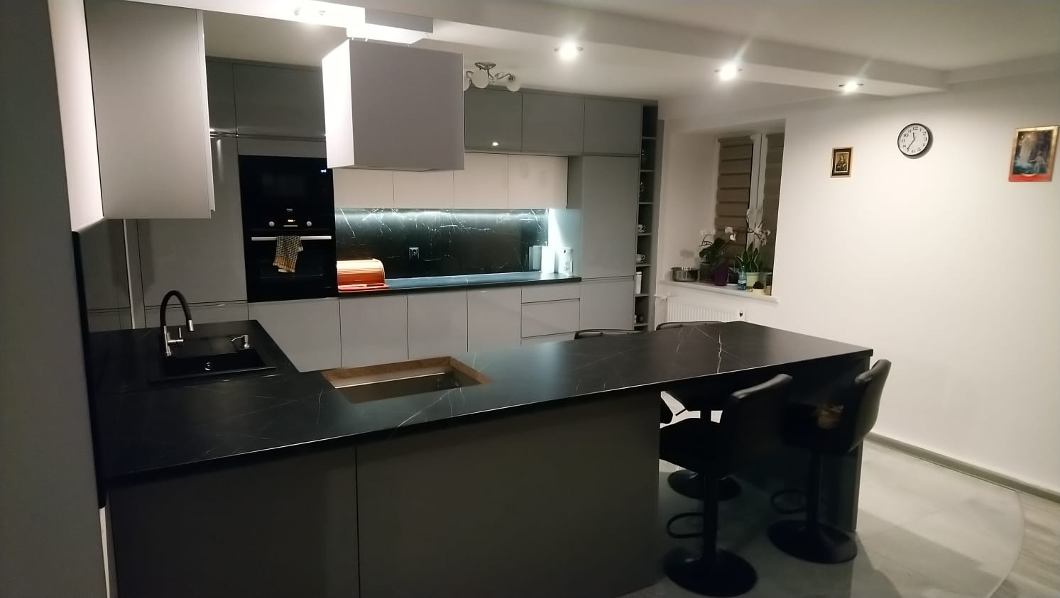 Nowoczesna kuchnia z wyspą i czarnym blatem imitującym marmur, szare szafki, oświetlenie LED pod szafkami, widok na okno z roślinami.