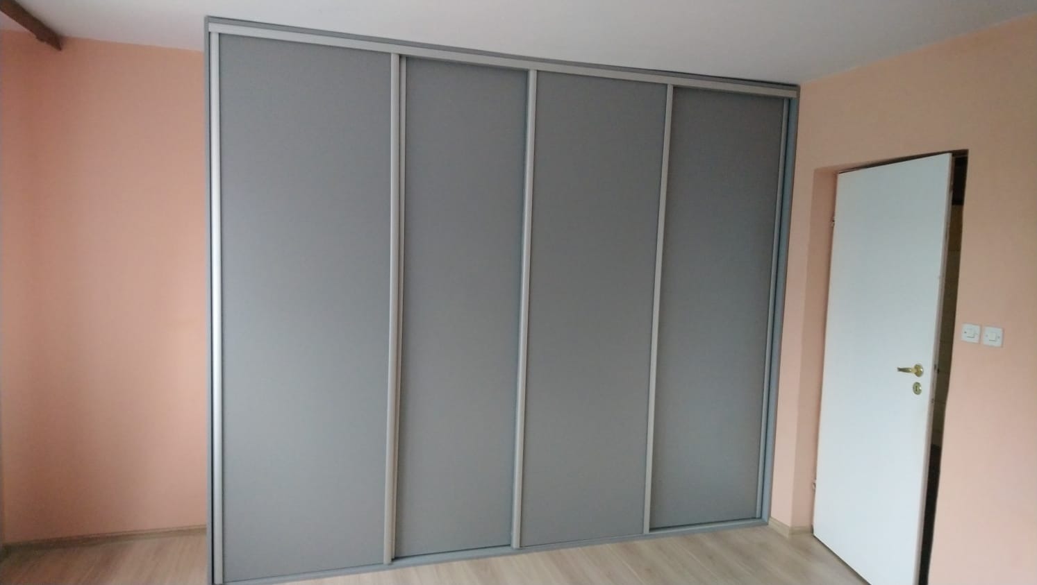 Czterodrzwiowa szafa wnękowa z przesuwnymi, szarymi drzwiami w pokoju o ścianach w kolorze brzoskwiniowym, widoczna biała ościeżnica drzwiowa po prawej stronie.