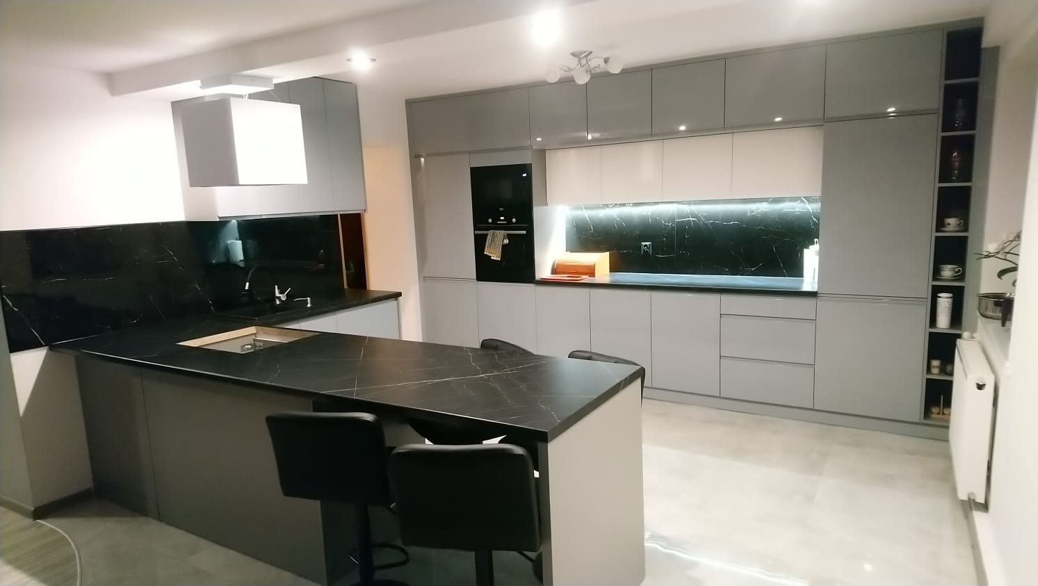 Nowoczesna kuchnia w odcieniach szarości z czarnym blatem marmurowym na wyspie i ścianie nad blatem, oświetlenie LED pod szafkami wiszącymi, czarne hokery przy wyspie.