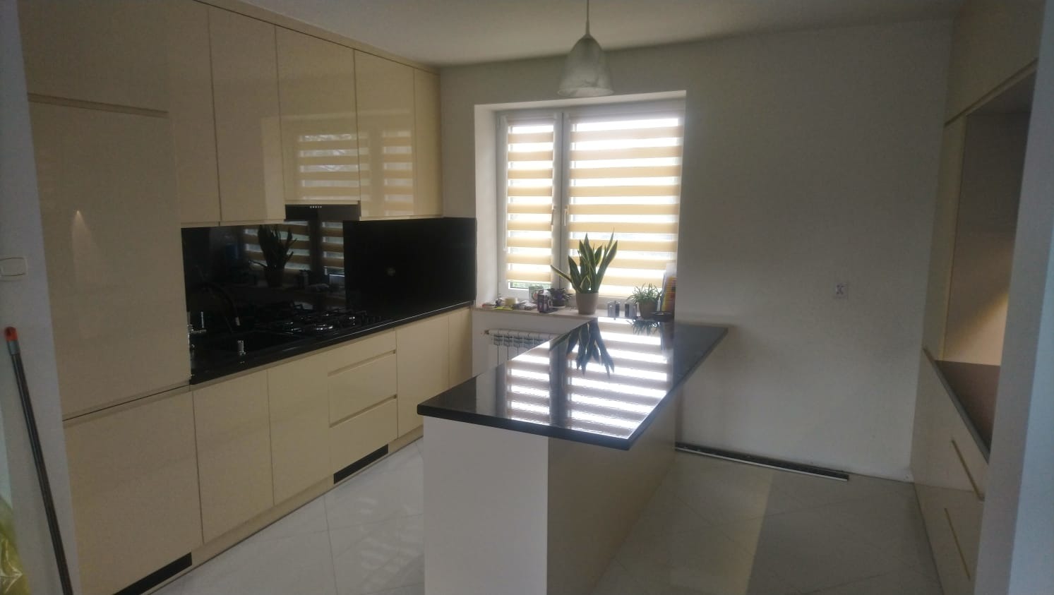 Jasna kuchnia z beżowymi frontami, czarnym blatem i wyspą kuchenną, rolety w oknie, odbicia światła na blacie.