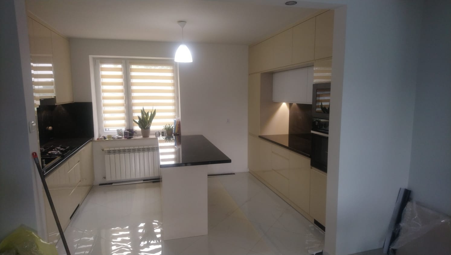 Jasna kuchnia z czarnym blatem, oświetleniem punktowym i wyspą kuchenną, widok na okno z roletami i roślinami doniczkowymi.