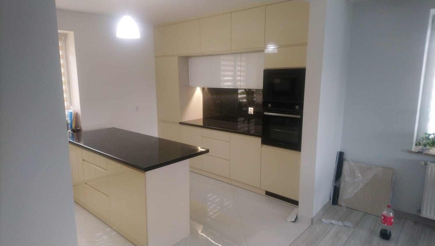 Beżowa kuchnia na wymiar z czarnym blatem i wyspą kuchenną, widoczne podłączenia elektryczne, biała podłoga z połyskiem.