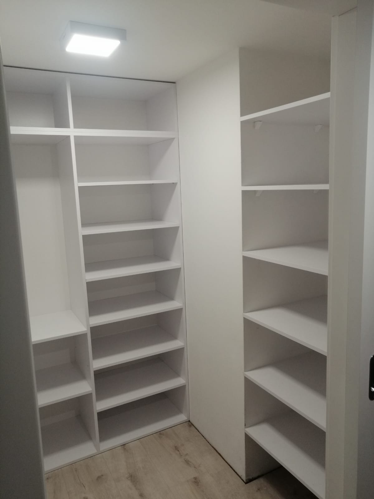 Wnętrze garderoby z dwiema białymi szafami wnękowymi z półkami, oświetlenie sufitowe, jasna podłoga imitująca drewno.