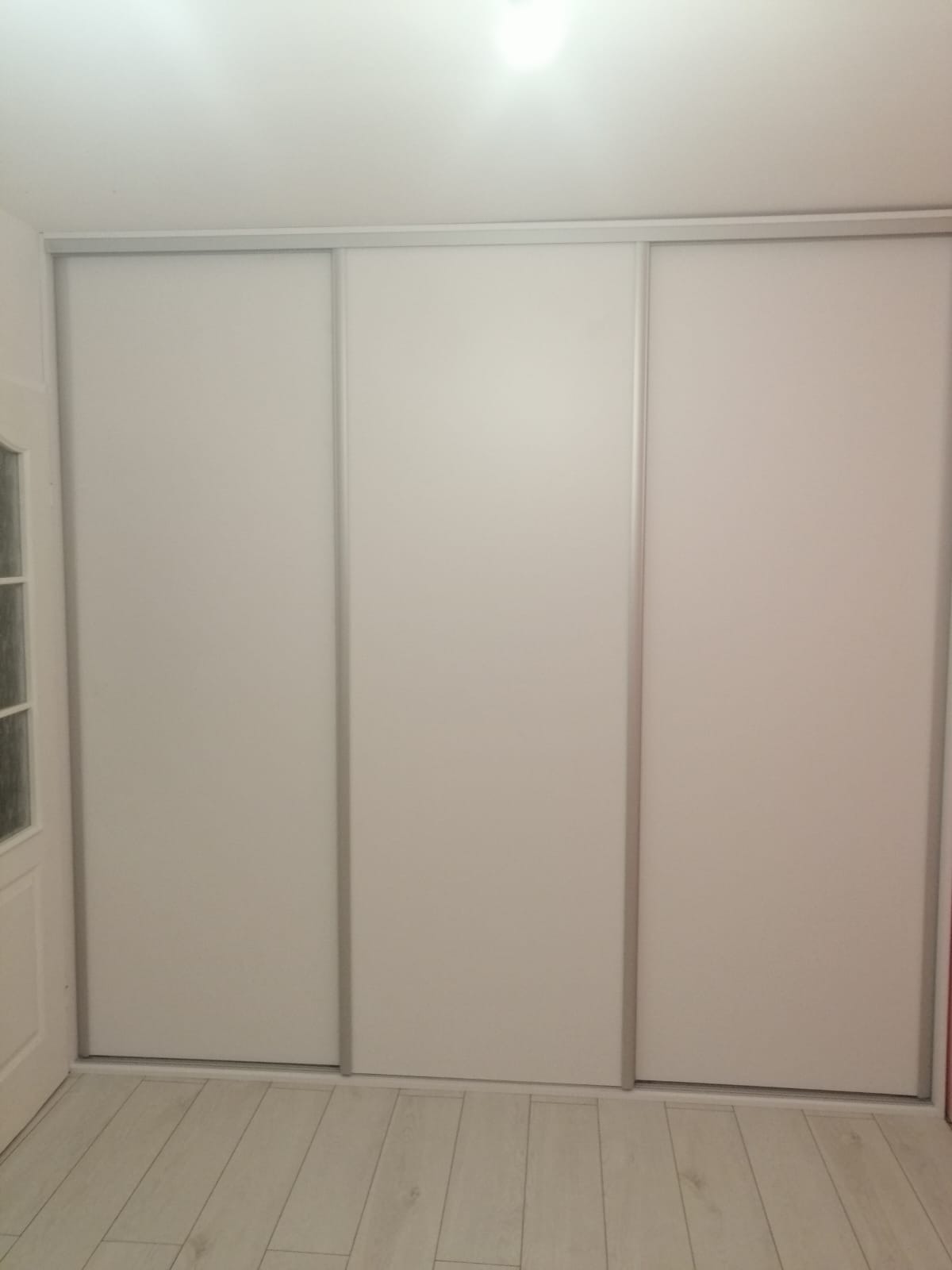 Trzydrzwiowa, biała szafa wnękowa z przesuwnymi drzwiami i srebrnymi prowadnicami, widoczna na tle jasnej podłogi i ściany, z fragmentem białych drzwi po lewej stronie.