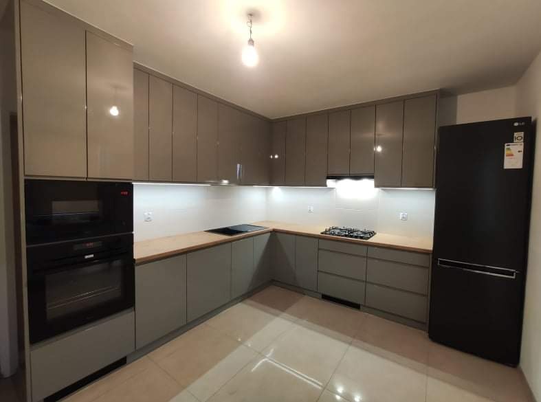 Nowoczesna, szara kuchnia z błyszczącymi frontami szafek, drewnianym blatem, wbudowanym piekarnikiem i czarną lodówką LG.