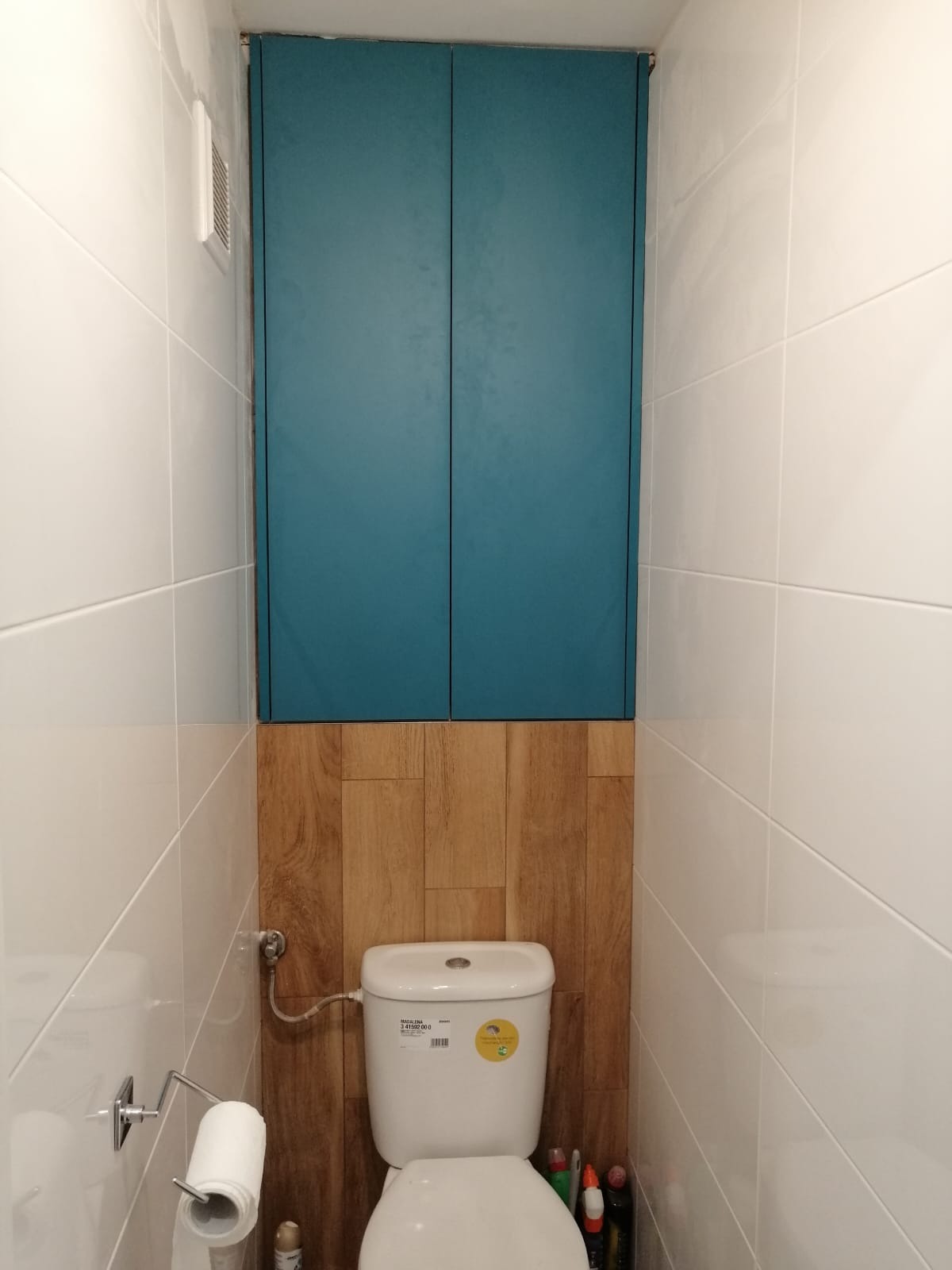 Wnętrze toalety z zabudowaną szafką w kolorze turkusowym nad deską sedesową, wykończoną drewnopodobnymi panelami, oraz białą glazurą na ścianach.