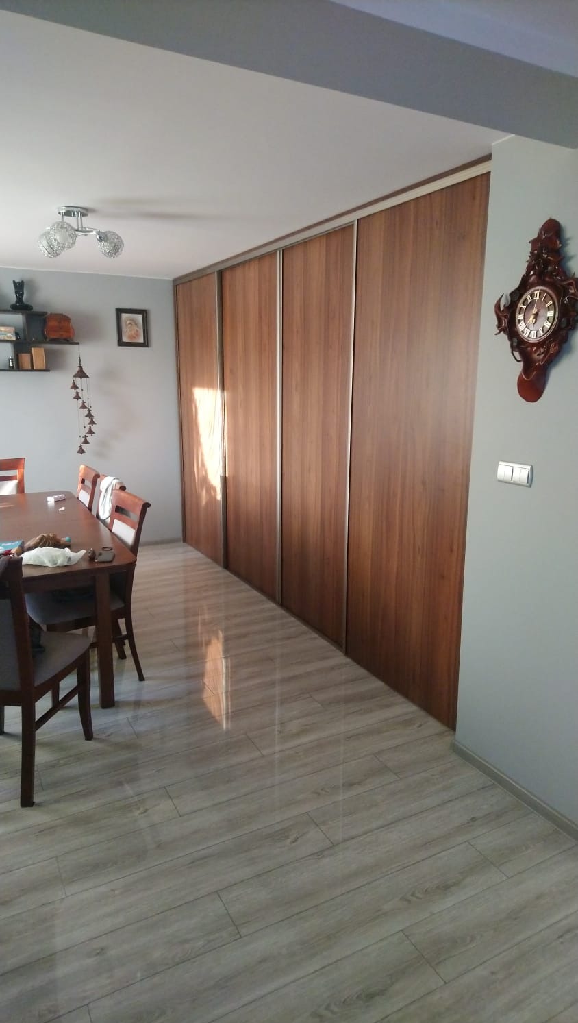 Długi rząd szaf wnękowych z przesuwnymi drzwiami o drewnianym wykończeniu w jadalni, widoczny stół z krzesłami i dekoracyjny zegar na ścianie.