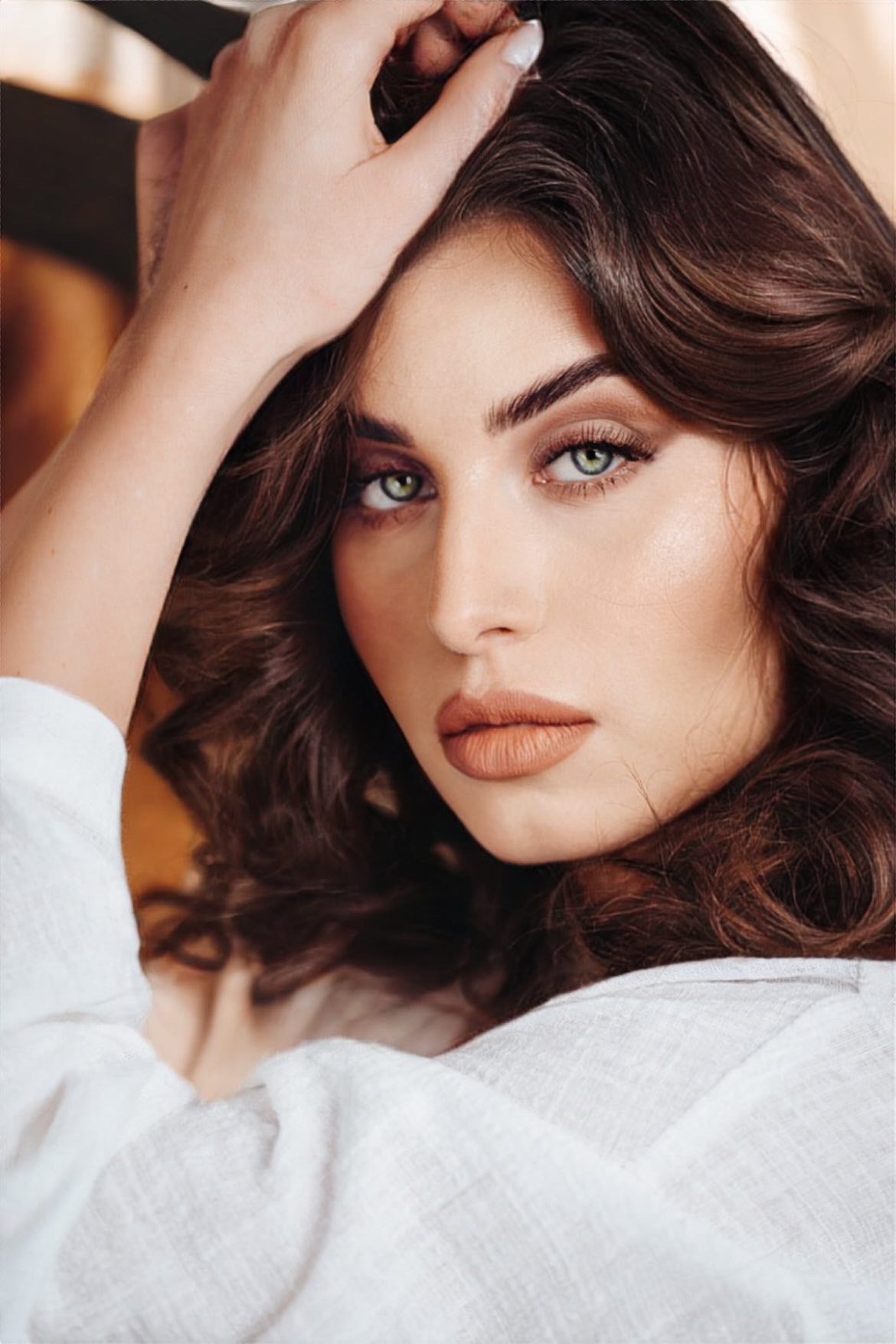 Portret kobiety z falowanymi, brązowymi włosami, delikatnym makijażem w odcieniach nude i zielonymi oczami, dłoń uniesiona do góry, biała koszula.