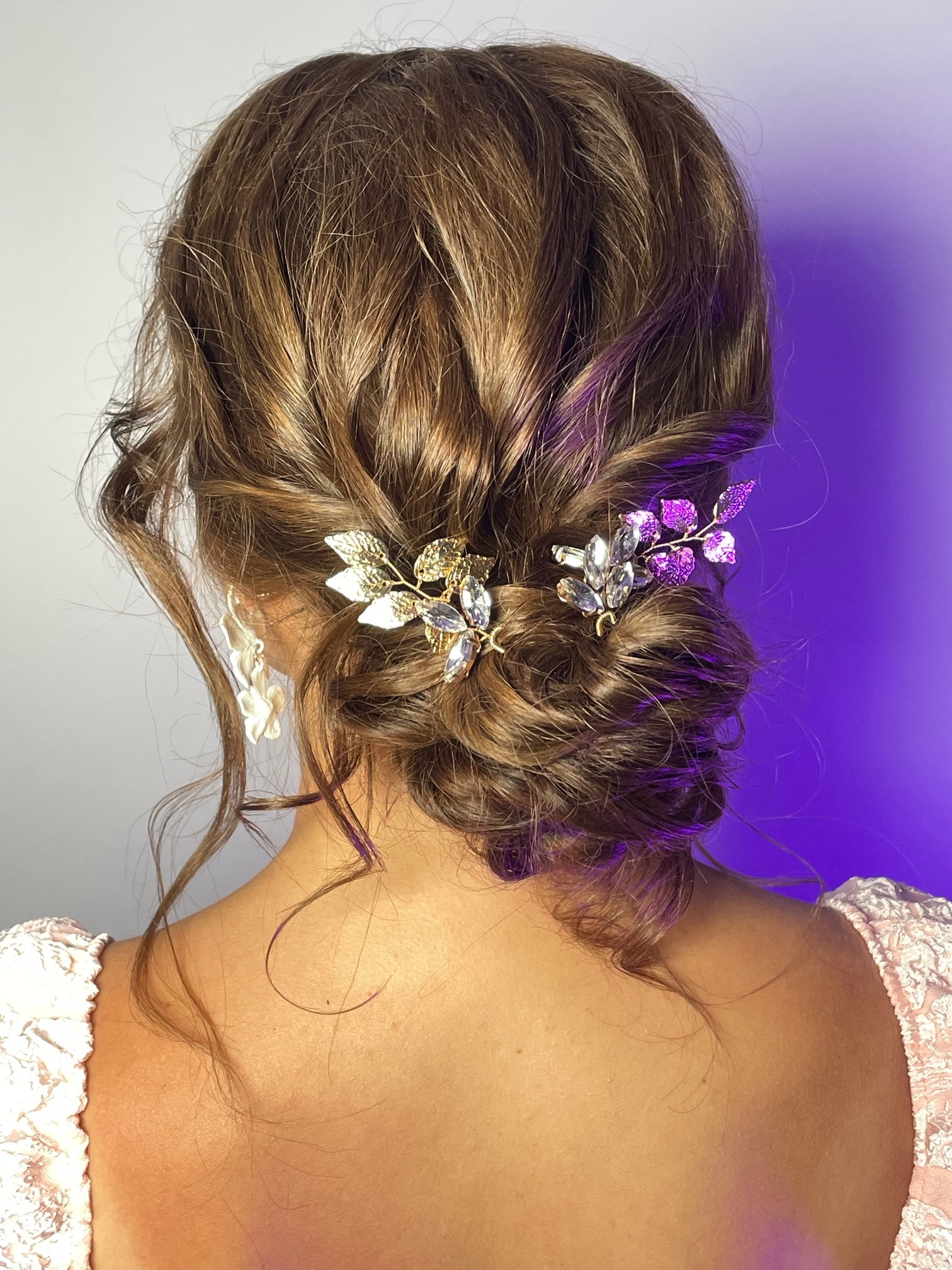Upięcie włosów z ozdobnymi spinkami w kształcie listków, złote i srebrne, na brązowych, kręconych włosach, widok z tyłu, oświetlenie fioletowe.
