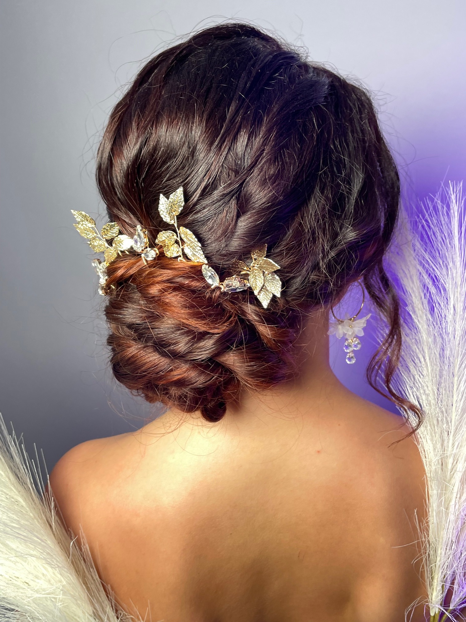 Upięcie włosów w kok z wplecioną ozdobą z liści i kryształków, widok z tyłu, uzupełnione kolczykami z kwiatami i piórami.