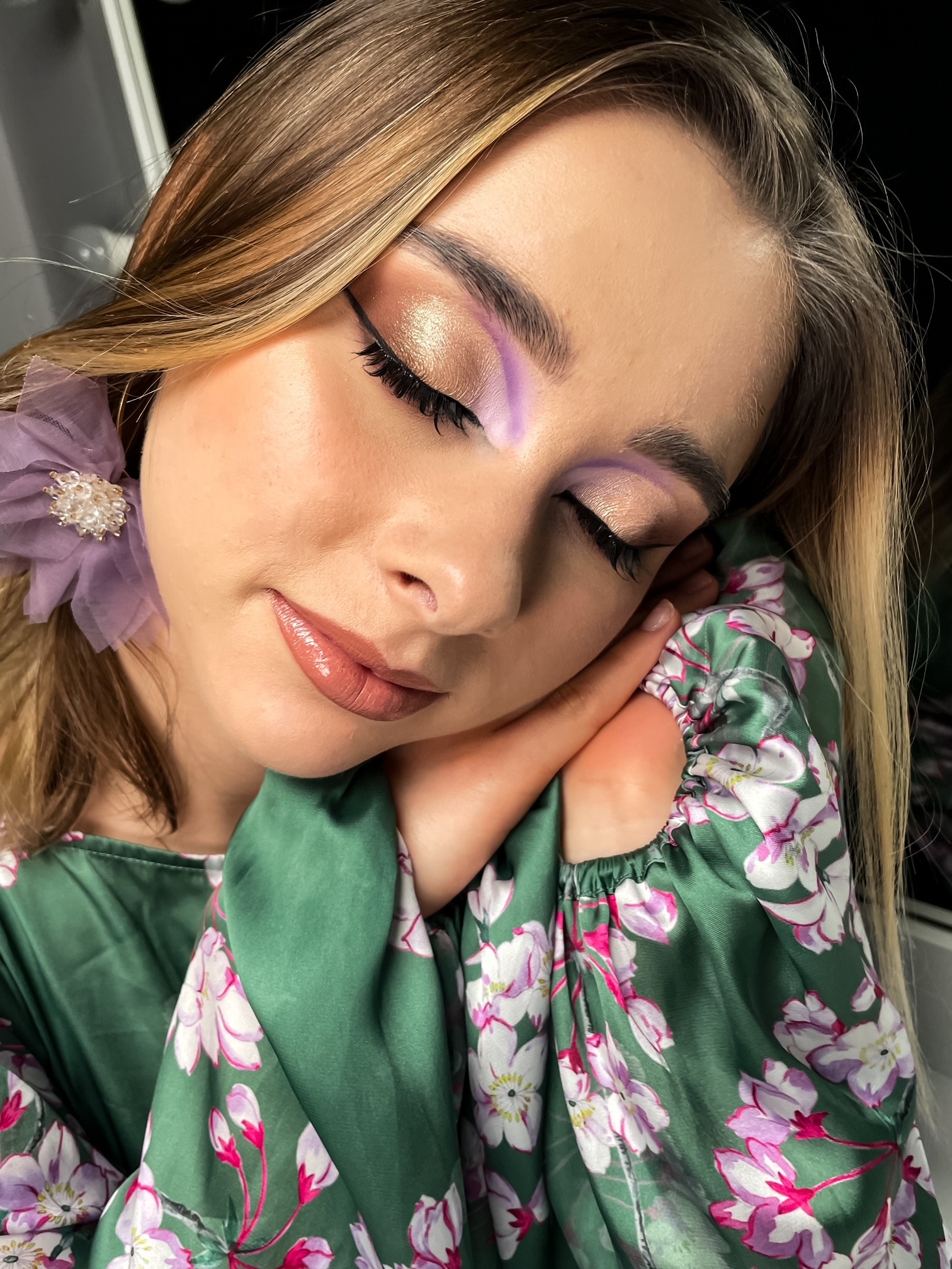 Portret kobiety z zamkniętymi oczami, prezentującej złoto-fioletowy makijaż oczu i błyszczące, brzoskwiniowe usta, z delikatnym kwiatem we włosach, w zielonej kwiecistej bluzce.